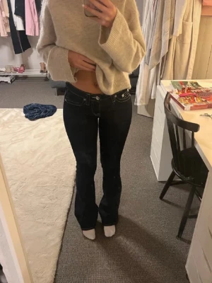 Mörkblå bootcut jeans True Religion - Säljer ett par mörkblå bootcut jeans från True Religion med kontrastsömmar och ikoniska fickdetaljer bak. Jeansen har låg midja och klassisk femficksdesign. Perfekta för dig som gillar en tidlös och trendig look.