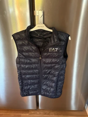 Mörkblå dunväst från EA7 Emporio Armani - Snygg mörkblå dunväst från EA7 Emporio Armani med quiltad design och dragkedja framtill. Västen har hög krage och logga på bröstet. Perfekt för lager-på-lager och ger en sportig vibe till din outfit.