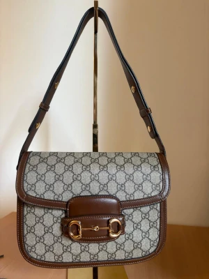 Gucci GG Supreme axelremsväska brun - Snygg axelremsväska från Gucci i GG Supreme canvas med bruna läderdetaljer och guldfärgad hästbitsdetalj framtill. Väskan har justerbar rem och klassiskt monogrammönster. Perfekt för dig som gillar lyxig och tidlös design.