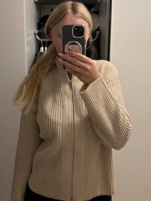 Beige ribbstickad zip cardigan Lager 157  - Beige ribbstickad zip cardigan från 157 med hög krage och dragkedja framtill. Tröjan har en avslappnad passform och långa ärmar, perfekt för lager-på-lager. Enkel och stilren design som passar till det mesta.