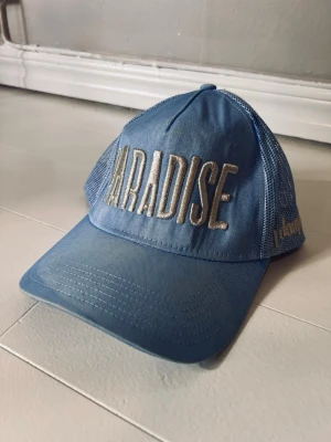 Blå keps  PARADISE- j dauphin  - Snygg ljusblå truckerkeps med stort broderat PARADISE-tryck framtill och mesh på sidorna. På sidan finns även broderad text 'dauphin' i beige. Perfekt för en chill och trendig look.