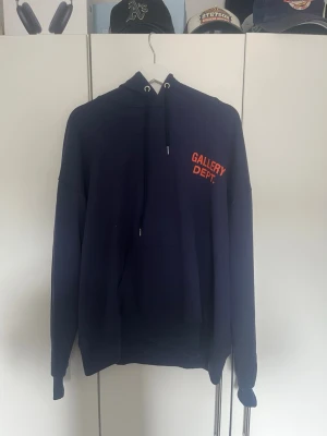 Mörkblå hoodie Gallery Dept. - Snygg mörkblå hoodie från Gallery Dept. med orange tryck fram och bak. Klassisk huva med dragsko och stor magficka. Oversized passform och ribbade muddar. Perfekt för dig som gillar streetwear och vill sticka ut med tydlig logga.