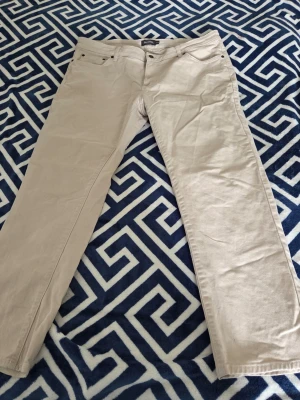 Beiga raka jeans - Säljer ett par beiga jeans med rak passform och klassisk femficksdesign. Jeansen har bälteshällor och knappgylf. Perfekta för dig som vill ha ett par ljusa och stilrena byxor till garderoben.