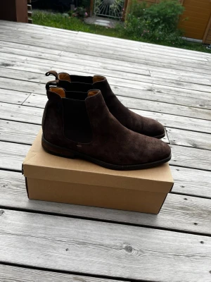 Mörkbruna Chelsea boots i mocka - Storlek: 44👌🏼Riktigt Schyssta chelsea boots som kan användas både vardagligt men även till mer uppklädda tillfällen. Mörkbrun mocka med klassisk siluett och relativt god kvalitét. Låg klack, och relativt smal och nätt passform. (Inte den breda käng varianten). Hör av dig vid frågor eller behov av fler bilder!