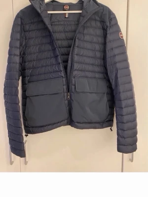 Mörkblå pufferjacka från Colmar - Snygg mörkblå pufferjacka från Colmar med två stora fickor framtill och diskret logga på ärmen. Jackan har quiltad design, dragkedja och hög krage. Perfekt för kalla dagar och riktigt skön att bära. Materialet är lätt och isolerande.