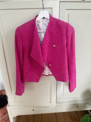Kort rosa kavaj med guldknappar, Morris - Säljer en kort, figursydd kavaj i starkt rosa med två dekorativa guldknappar framtill. Kavajen har en strukturerad, tweed-liknande yta och helfodrad insida. Perfekt för att lyfta vilken outfit som helst med färg och stil.