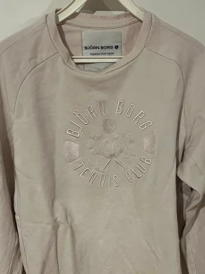 Ljusrosa sweatshirt från Björn Borg - Ljusrosa sweatshirt från Björn Borg med broderad text 'Tennis Club' och två tennisrack på bröstet. Tröjan har rund halsringning och långa ärmar. Perfekt för en sportig och avslappnad stil. Pris kan diskuteras vid snabb affär