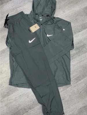 Nike miller - Snyggt mörkgrått träningsset från Nike med matchande jacka och byxor. Jackan har huva, dragkedja och vita Nike-loggor på bröstet och byxan. Materialet är lätt och andas, perfekt för träning eller uppvärmning. Elastisk midja på byxorna för skön passform.