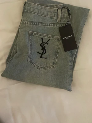 Jeans med ysl logga  - Ljusblå jeans i klassisk rak modell. Jeansen har en bakficka med stor svart YSL-logga broderad och är tillverkade i denim. Perfekta för dig som vill ha en stilren och exklusiv look.