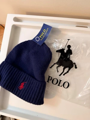 Mörkblå ribbstickad mössa Polo Ralph Lauren - Mörkblå ribbstickad mössa från Polo Ralph Lauren med klassisk uppvikt kant och röd broderad logotyp framtill. Perfekt accessoar för kyliga dagar och enkel att matcha med olika outfits.