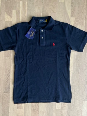 Ralph Lauren navy polo shirt size M - Brand new, never worn Polo Ralph Lauren Custom Slim Fit polo shirt in classic navy blue. Comes with original tags attached and original packaging.  * Condition: Brand New with Tags (NWT) * Color: Navy Blue * Fit: Custom Slim Fit * Size: M * Material: 100% Cotton * No flaws, stains, holes, or signs of wear. * Authentic Polo Ralph Lauren. * Retail ≈ 180$ A timeless wardrobe essential, perfect for both casual and smart-casual outfits. 