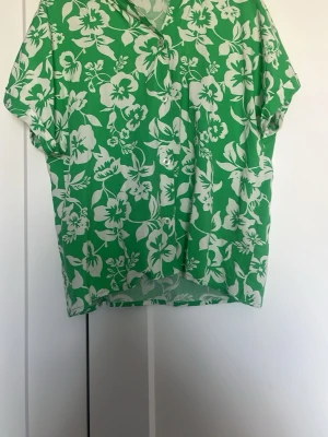 Grön blommig kortärmad skjorta - Snygg grön skjorta med vitt blommigt mönster. Skjortan har korta ärmar, knappar framtill och en avslappnad passform. Perfekt för dig som vill sticka ut med en färgglad och somrig stil.