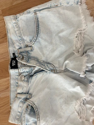 Ljusblåa jeansshorts med slitningar - Säljer ett par ljusblåa jeansshorts med råa, slitna kanter och fransiga detaljer. Shortsen har klassisk femficksdesign, bälteshällor och knappgylf. Perfekta för varma dagar och en avslappnad stil.