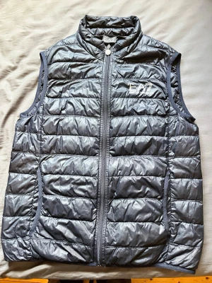 Mörkblå dunväst från EA7 Emporio Armani - Snygg mörkblå dunväst från EA7 Emporio Armani med quiltad design och hög krage. Västen har dragkedja framtill, två sidofickor och logga på bröstet. Perfekt för lager-på-lager och ger en sportig look.