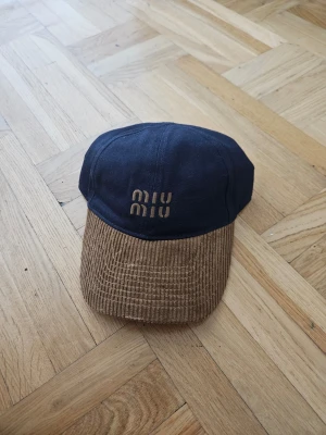 Miu Miu keps i blått och brunt - Snygg keps från Miu Miu med mörkblå ovandel i tyg och brunt skärm i manchestertyg. Broderad Miu Miu-logga framtill i beige. Perfekt accessoar för en trendig och avslappnad stil.