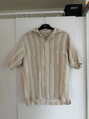 Beige randig skjorta från Yoke - Snygg beige skjorta från Yoke med vertikala ränder i ljusa toner. Skjortan har korta ärmar, klassisk krage, bröstficka och knäppning framtill. Perfekt för en avslappnad och stilren look.