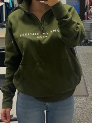 Olivgrön hoodie från Tommy Hilfiger - Snygg olivgrön hoodie från Tommy Hilfiger med halv dragkedja och stor magficka. Tryckt logga och flagga på bröstet. Mjuk insida och ribbade muddar vid ärmar och midja. Perfekt för en chill och avslappnad stil.