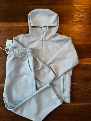 Nike NOCTA ljusblå tracksuit set - Nike x NOCTA Tracksuit i mycket fint skick. Tröjan är använd endast 4 gånger och byxorna är helt nya. Säljs då den tyvärr inte passar mig. Storlek XL, men upplevs  mindre i passformen. Är mer är M/L.  Säljs till ett bra pris då är behöver göra plats i garderoben.