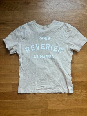 Beige t-shirt med tryck - Säljer en beige t-shirt med texten 'PARIS RÊVERIES LE MARAIS' i vitt framtill. T-shirten har rund halsringning och korta ärmar. Tillverkad i mjuk bomull, perfekt för en avslappnad och trendig look.