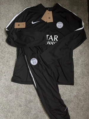 PSGTracksuit - Säljer ett svart träningsset från PSG och Nike med långärmad tröja och matchande byxor. Setet har vita detaljer längs ärmar och ben, PSG-logga på bröst och byxa samt Nike swoosh. Materialet är lätt och andas, perfekt för träning eller uppvärmning.
