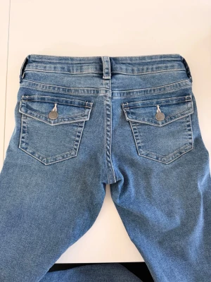 Blåa bootcut  jeans med ficklock - Snygga blåa bootcut  jeans i klassisk denim med ficklock och silverknappar bak. Jeansen har normalhög midja, bälteshällor och femficksmodell. Materialet är mjukt och stretchigt för en bekväm passform. Perfekta till en avslappnad look.