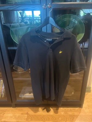 Svart pikétröja från Lyle & Scott - Klassisk grå/svart pikétröja från Lyle & Scott med korta ärmar och gul broderad logga på bröstet. Tröjan har krage och knappar vid halsen, perfekt för en clean och stilren look. PRUTA