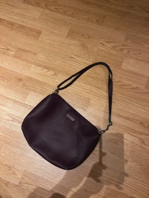 Mörklila axelväska från Guess - Snygg mörklila axelväska från Guess i ett stilrent, mjukt konstläder med struktur. Väskan har justerbar axelrem och silverfärgade metalldetaljer. Perfekt storlek för det viktigaste och enkel att matcha till olika outfits.