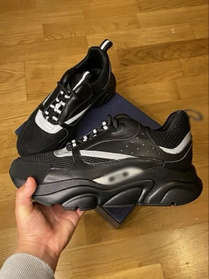 Chunky svarta sneakers med vita detaljer - Säljer ett par chunky svarta sneakers med vita detaljer och meshpaneler. Skorna har en kraftig sula, svart ovandel i läder och mesh samt vita inslag på sidan och snörningen. Perfekta för dig som gillar streetwear och en sportig look.