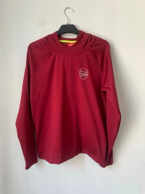 Röd Arsenal hoodie med huva - Snygg röd hoodie med Arsenal-logga på bröstet. Tröjan har långa ärmar, huva och är gjord i ett mjukt, sportigt material som passar perfekt till träning eller chill. Enkel design med klubbens emblem tryckt i vitt framtill. Storleken är 12-13 år men den är stor och passar för lite äldre typ 15 år.