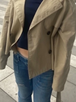Kort beige trenchcoat med knappar - Trendig kort trenchcoat i beige med dubbelknäppning och bred krage. Jackan har en avslappnad passform och långa ärmar. Perfekt för lager på lager och streetstyle. Tillverkad i ett lätt tyg som ger en snygg siluett.