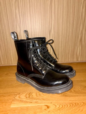 Svarta lackade Dr. Martens-kängor med snörning - Säljer ett par svarta, glansiga Dr. Martens-kängor i lackmaterial med grov sula och snörning framtill. — Kängorna har dragflikar bak med text och metallhål för snören. — Perfekta för dig som vill ha en edgy och stilren look. — Sparsamt använda, i mycket bra skick. — Skokartongen saknas tyvärr. — Nypris ca. 2 200 kr.