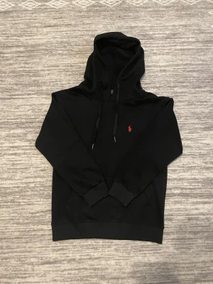 Svart Polo Ralph Lauren hoodie  - Snygg svart hoodie från Polo Ralph Lauren med klassisk röd logga broderad på bröstet. Hoodien har huva med dragsko, känguruficka och ribbade muddar vid ärmslut och nederkant. Perfekt för en avslappnad och stilren look. Lite liten i storleken  passar för xs oxå 