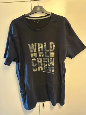 Svart t-shirt från Firefly med tryck - Svart t-shirt från Firefly i storlek XXL med coolt tryck på bröstet där det står 'WRLD CREW' i ett mönster med camouflage. Klassisk rund halsringning och korta ärmar. Perfekt för en avslappnad och trendig stil.