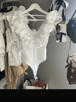 Zara body - Säljer en vit body med  V-ringning och stora volangdetaljer runt halsen och axlarna. Bodyn har en feminin och trendig look, är ärmlös och tillverkad i ett mjukt, stretchigt material. Perfekt för dig som vill ha något extra till outfiten.