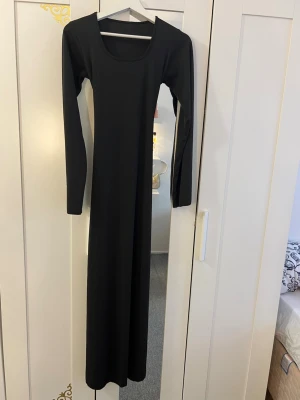 Svart långklänning med snörning - Stilren svart långklänning med lång ärm och rundad urringning. Klänningen har snygg snörningsdetalj i ryggen som ger en extra touch. Passformen är kroppsnära och materialet ser ut att vara mjukt och stretchigt, perfekt för en trendig look.