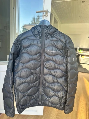 Svart peak dunjacka - Säljer en svart dunjacka från Peak Performance med huva och hel dragkedja framtill. Jackan har ett vågformat quiltat mönster och är lätt vadderad för extra värme. Perfekt för kalla vinterdagar och riktigt snygg till streetwear-stilen. Den har två lagningar på arm och rygg 