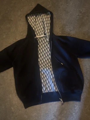 Blå d1or zip  - Blå d1or zip. Insidan av huvan har ett ikoniskt D1or-mönster i svart och vitt. Tröjan har fickor framtill och ribbade muddar. Perfekt för dig som gillar exklusiva streetwear-plagg med lyxig känsla.