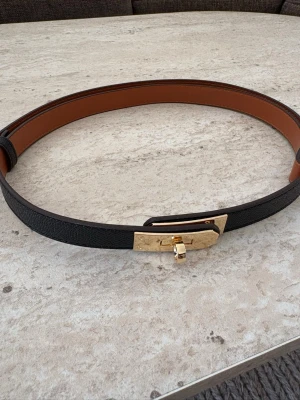 Svart bälte med guldfärgat spänne - Snyggt svart Kelly bälte i skinn med guldfärgat, rektangulärt spänne och brunt foder på insidan. Perfekt accessoar för att lyfta din outfit med en elegant touch. Storleken går att justera