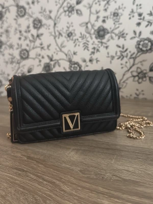  Victoria Secret Svart kuvertväska med guldkedja - Snygg svart kuvertväska i quiltat läder med guldfärgad kedja och V-detalj på locket. Väskan har flera fack inuti och stängs med magnetlås. Perfekt för att bära det viktigaste och ger en lyxig känsla till din outfit.