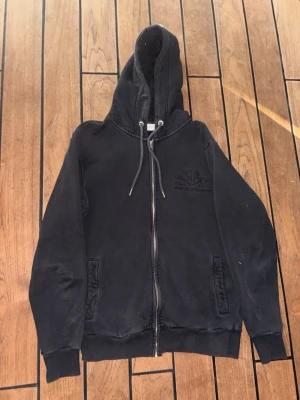 Svart zip hoodie med dragsko - Snygg svart hoodie med dragkedja och huva med dragsko. Två fickor framtill och ribbade muddar vid ärmar och nederkant. Diskret tryck på bröstet. Perfekt för en avslappnad och cool stil.