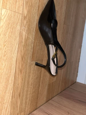 Svarta lackpumps med smal klack - Säljer ett par svarta pumps i glansigt lackmaterial med spetsig tå och smal klack. Skorna har öppen häl och justerbar rem med guldfärgat spänne. Perfekta för dig som gillar klassisk och elegant stil.