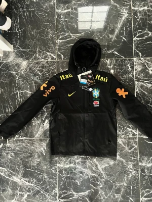 Svart Brasilien vindjacka med huva - Snygg svart vindjacka med huva och dragkedja, prydd med Brasilien fotbollslandslagets logga och flera sponsorloggor i gult och orange. Jackan har två fickor framtill och tryck på både bröst och ärmar. Perfekt för dig som gillar sportig stil.