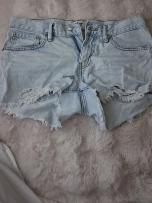 Ljusblå slitna jeansshorts - Säljer ett par ljusblå jeansshorts med råa, fransiga kanter och slitningar framtill. Klassisk femficksmodell med knappgylf och bälteshällor. Perfekta för sommaren och ger en avslappnad vibe.