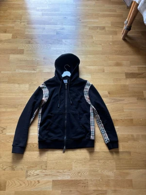 Burberry Nova Check Zip Hoodie -   Stilren och exklusiv Burberry-hoodie med ikoniska Nova Check-paneler längs sidorna och axlarna. Tillverkad i mjuk premiumbomull och utrustad med tvåvägsdragkedja, huva och framfickor.  • Mycket fint skick • Klassisk Burberry-design • Bekväm passform • Nypris ca 8 000–10 000 kr  En eftertraktad modell som är svår att hitta. Perfekt för både vardag och en mer uppklädd look. Snabb frakt vid smidig affär. Ända är en liten lös stitch på necktagen.