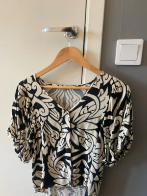 Svartvit blus med puffärm och mönster - Unik svartvit blus med stora puffärmar och v-ringning. Blusen har ett iögonfallande grafiskt mönster och smockad nederkant som ger en snygg passform. Perfekt för dig som vill sticka ut med din stil. Köpt på en affär vid namn fabriken, vet ej vad de är för märke. Aldrig använd
