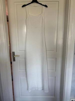 Vit lång stickad klänning - Säljer en vit långklänning med ribbad struktur. Klänningen är stickad och har en rak, kroppsnära passform som ger en stilren och enkel look. Aldrig använd. Skulle inte säga att den är genomskinlig ifall du inte har svarta underkläder.