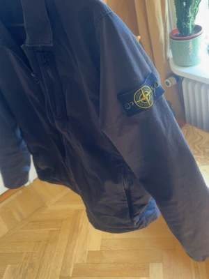 Svart jacka från Stone Island - Säljer en svart jacka från Stone Island med den klassiska logotypen på ärmen. Jackan har dragkedja framtill och en stilren, enkel design. Perfekt för dig som gillar streetwear och vill ha något som sticker ut.