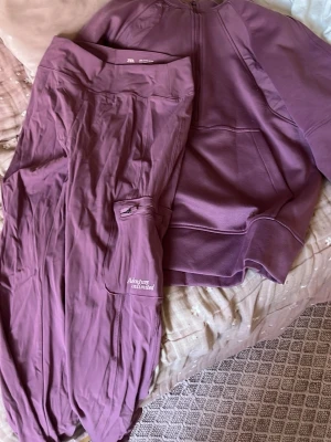 Lila loungewear set från Zara - Säljer ett lila loungewear set med ziptröja och matchande byxor. Tröjan har dragkedja och avslappnad passform, byxorna har ficka med texten 'Adventure unlimited' och resår i benslut. Perfekt för chill dagar hemma eller när du vill vara bekväm.