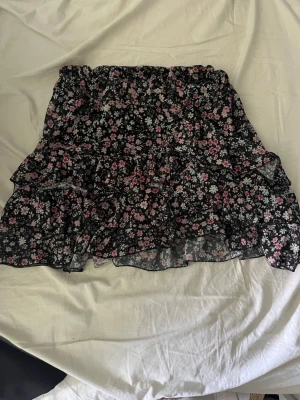 Svart blommig volangkjol - Söt kort kjol med svart botten och blommigt mönster i rosa och vitt. Kjolen har volanger och resår i midjan, vilket ger en luftig och flowy känsla. Perfekt för dig som gillar romantisk och feminin stil.