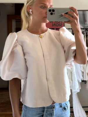 Ljus puffärmad blus med knappdetalj - Säljer en ljus, elegant blus med stora puffärmar och rund halsringning. Blusen har en dekorativ guldknapp vid halsen och är tillverkad i ett slätt, glansigt material som ger en exklusiv känsla. Något sliten längst ned på ena sidan.